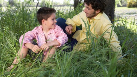 A father sits in the grass under a tree and talks with his son about executive function