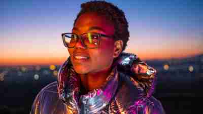 Young girl outdoors wearing eyeglasses and a shiny jacket that reflects colorful lights. Photo taken at disk, with city lights defocused at background.