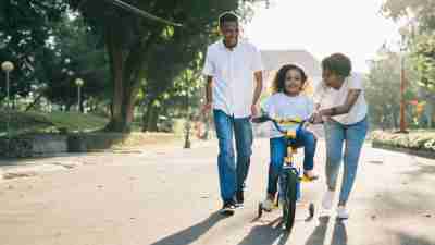 A mother and father smiling and teaching their child how to ride a bike. A supportive and encouraging parenting style known as authoritative parenting is best for neurodivergent children.
